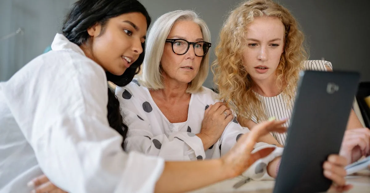 Groupe diversifié de femmes de plusieurs générations interagissant avec une tablette à l'intérieur.