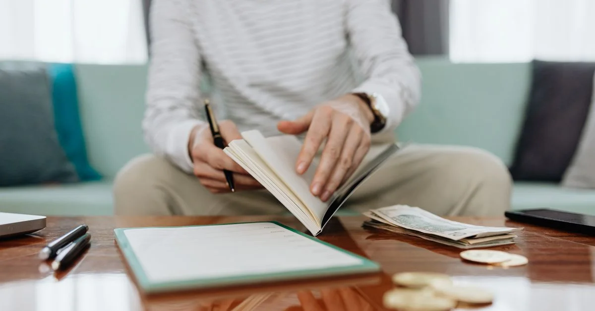 Individu examinant ses finances à une table avec un carnet, de l'argent et un ordinateur portable.