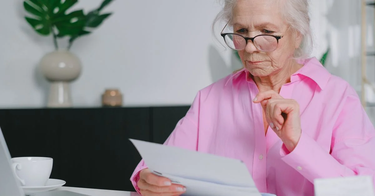 Femme âgée en chemisier rose lisant des documents dans son espace de travail à domicile.