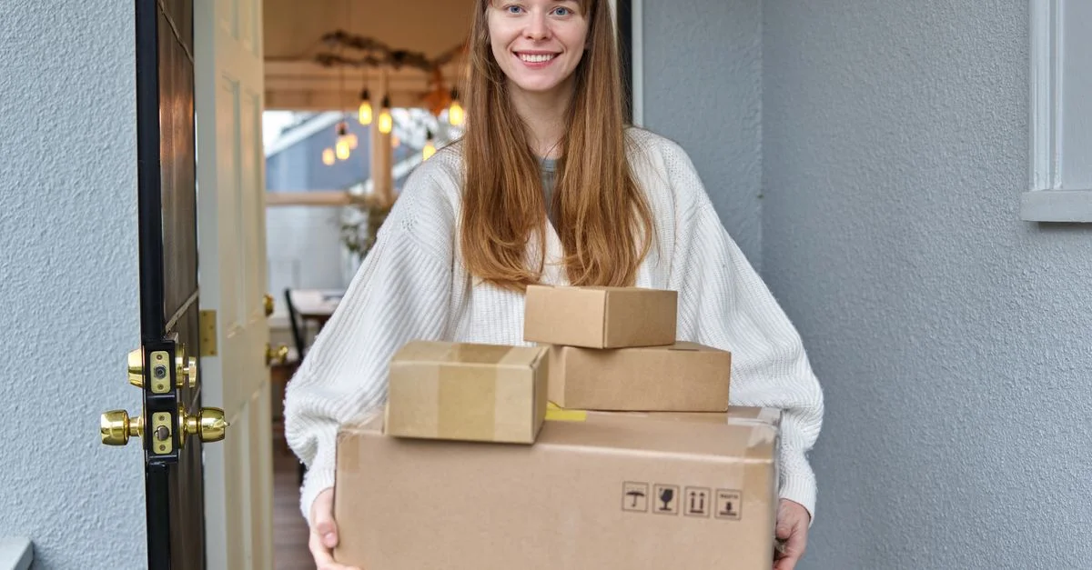 Une femme souriante tenant plusieurs cartons se tient dans une porte, accueillant chaleureusement.
