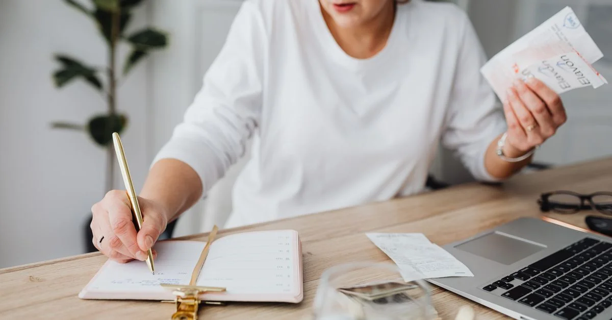 Femme gérant ses finances, écrivant sur un carnet et tenant des reçus à un bureau.