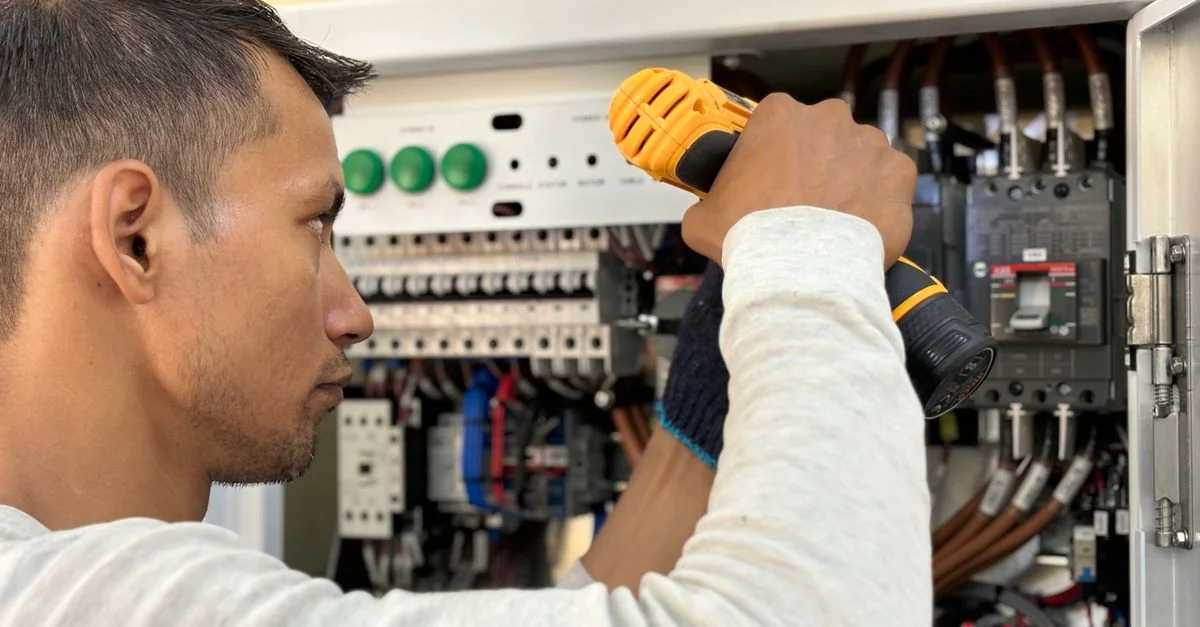 Électricien professionnel utilisant une perceuse sur un panneau de disjoncteurs intérieur.