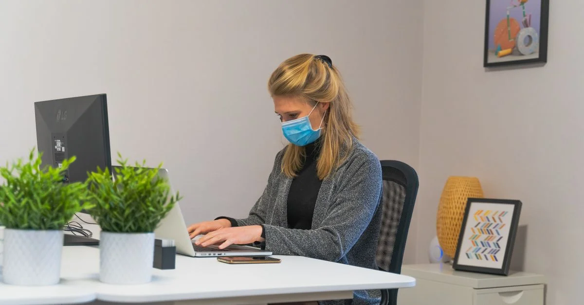Une femme portant un masque facial travaillant sur son ordinateur portable à un bureau de bureau intérieur.