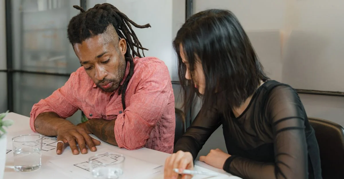Deux collègues collaborent sur des plans de conception dans un environnement de bureau moderne. Accent sur le travail d'équipe.