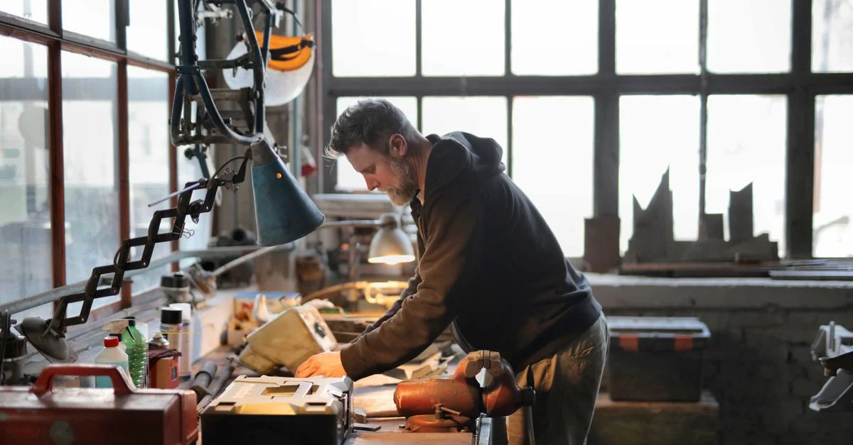 Un artisan dévoué travaillant intensément près d'une grande fenêtre dans un atelier ensoleillé rempli d'outils.