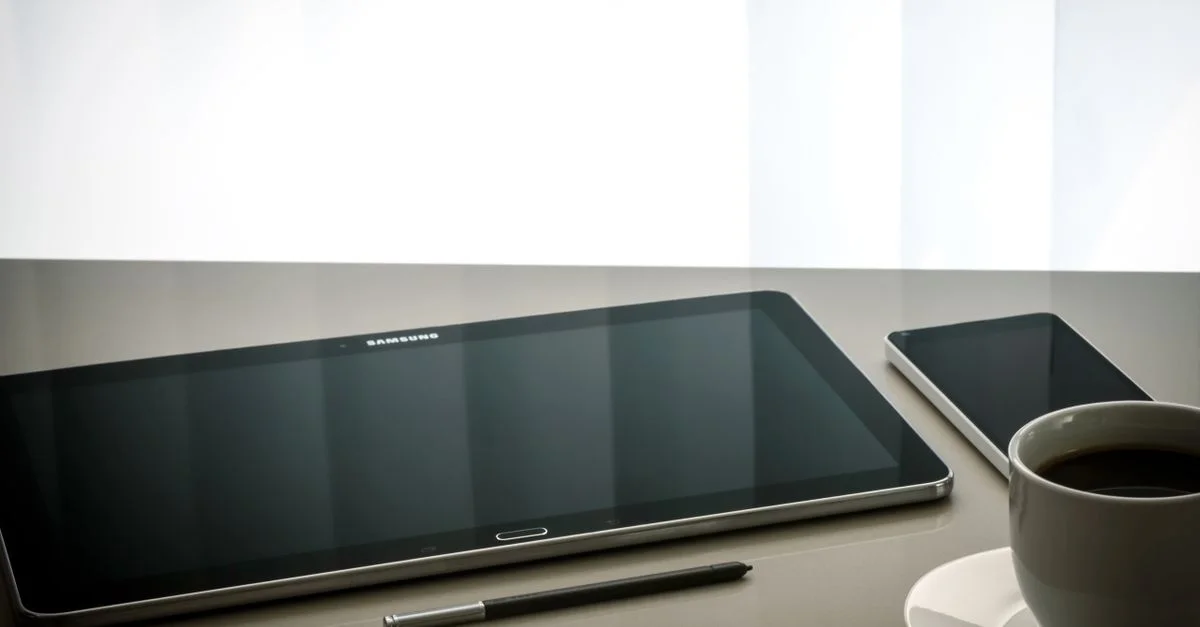 Configuration de bureau minimaliste avec une tablette, un smartphone, un stylo et une tasse de café sur une surface réfléchissante.