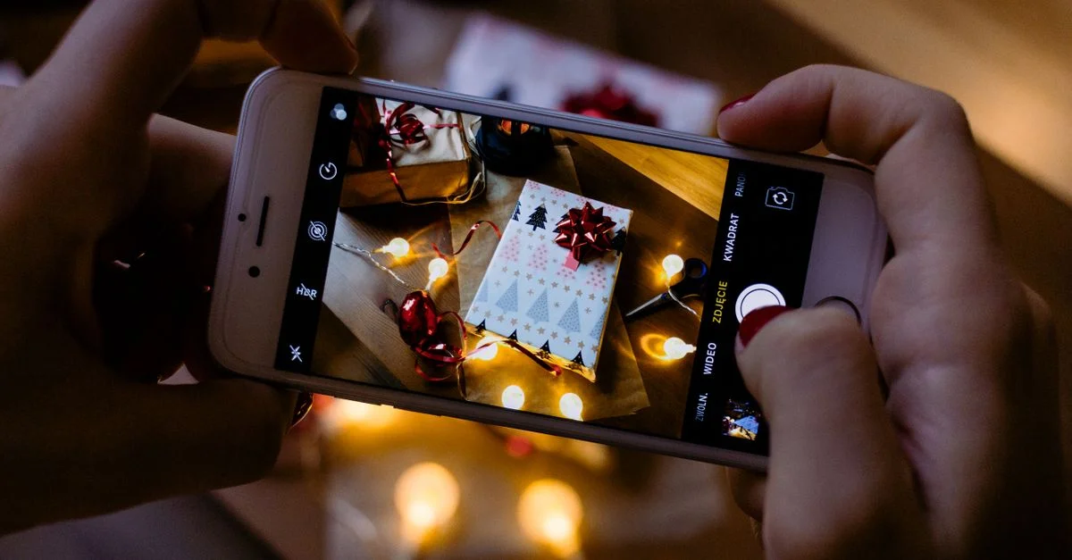 Mains tenant un smartphone capturant un cadeau de Noël avec des lumières festives.