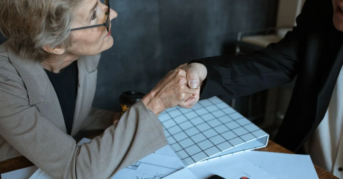 Femme senior en costume d'affaires serrant la main de l'autre côté d'une table dans un environnement de bureau.