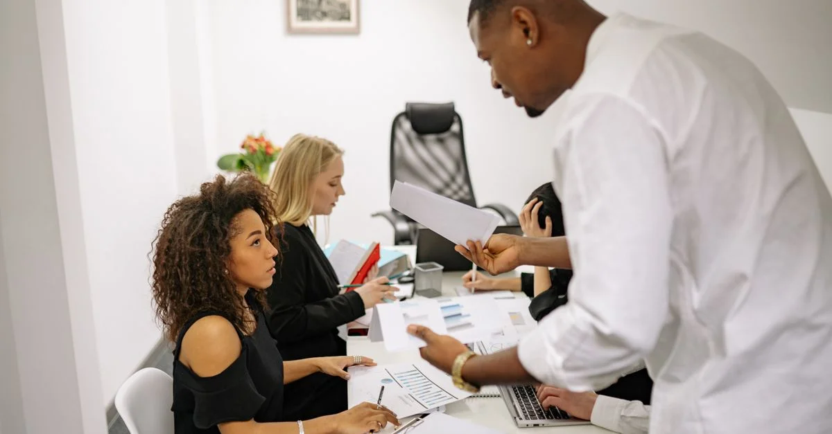 Une réunion de bureau où un homme discute avec une femme au sujet de documents, créant une tension.