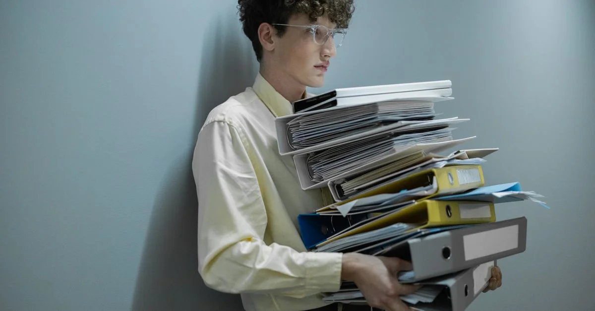 Un employé de bureau surchargé portant une grande pile de dossiers et de classeurs dans un environnement moderne.