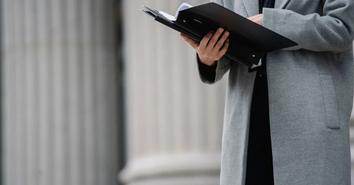 Femme professionnelle en manteau gris tenant un dossier de documents à l'extérieur d'un bâtiment avec des colonnes.