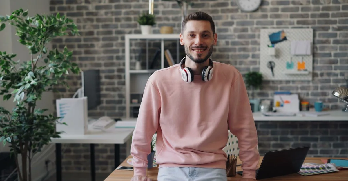 Homme habillé de manière décontractée souriant avec des écouteurs dans un cadre de bureau moderne, montrant un environnement de travail détendu.