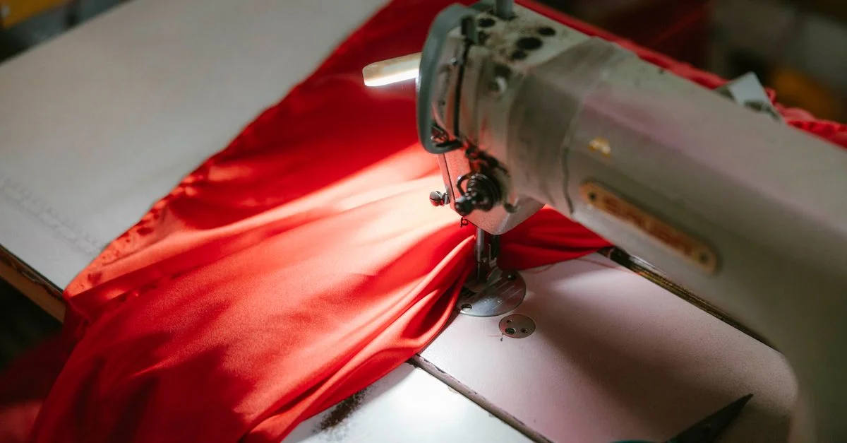 Gros plan sur une machine à coudre industrielle cousant un tissu rouge vif, éclairé sous un projecteur.
