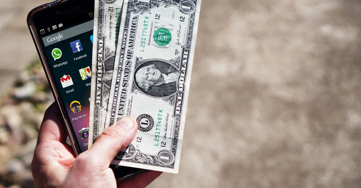 Gros plan sur une main tenant des billets en dollars américains et un smartphone en extérieur, mettant en avant la technologie financière.