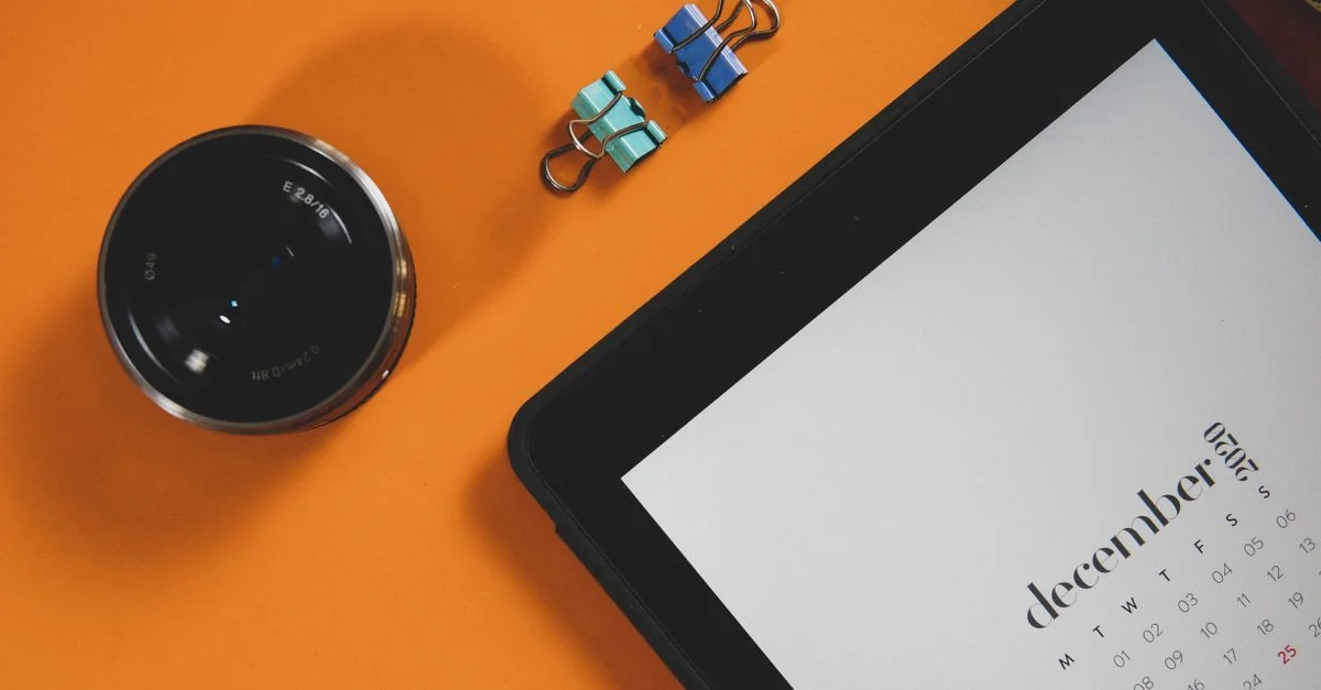 Un plan à plat d'un calendrier numérique, d'un objectif d'appareil photo et de trombones sur une surface orange.