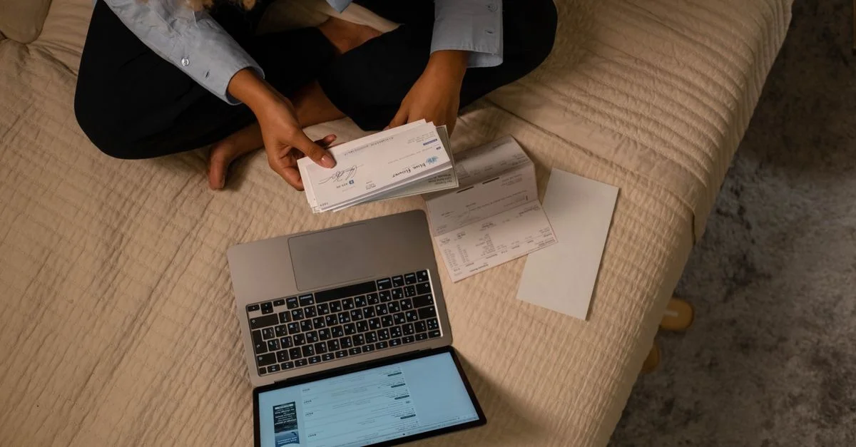 Vue en plongée d'une femme organisant ses finances sur un lit avec un ordinateur portable et des chèques.
