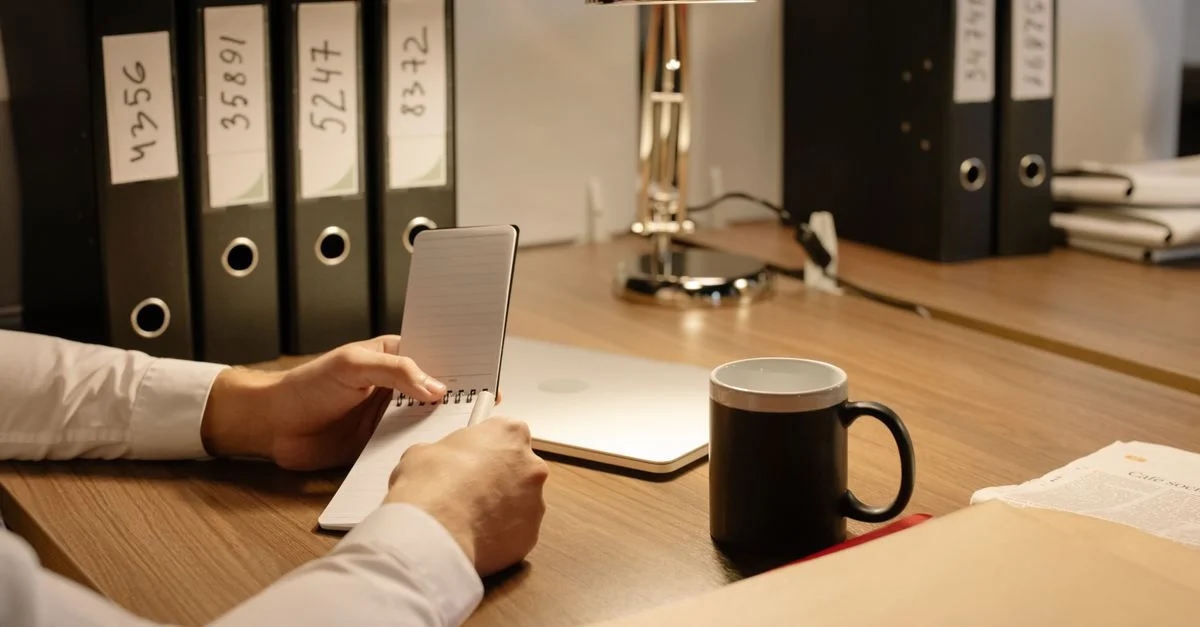 Bureau avec une personne tenant un carnet, une tasse de café, des dossiers et un ordinateur portable sur une table en bois.