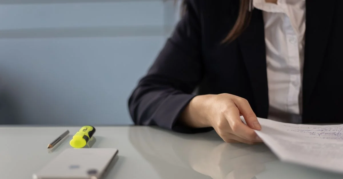 Professionnel de la business examinant des documents avec un surligneur et un téléphone sur un bureau en verre, idéal pour des scènes de bureau.