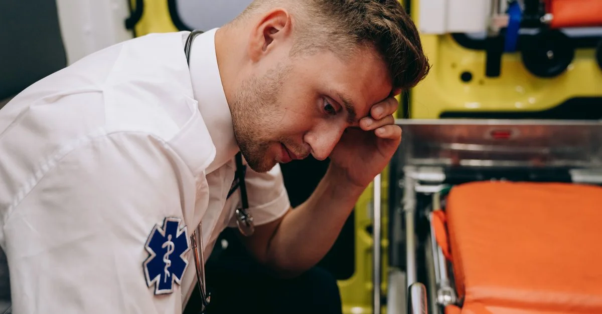 Un ambulancier fatigué assis dans une ambulance, la main sur la tête, indicatif de stress.