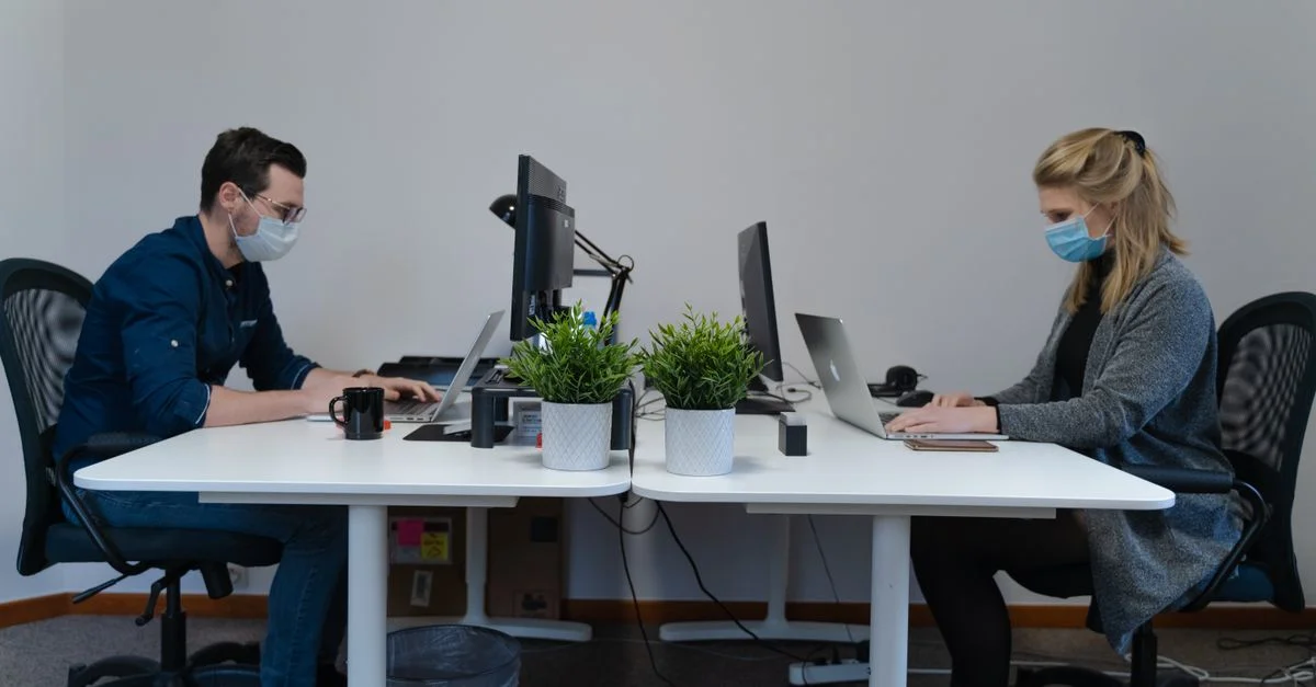 Deux employés travaillent dans un bureau avec des masques, séparés par des bureaux, utilisant des ordinateurs portables pour être productifs.