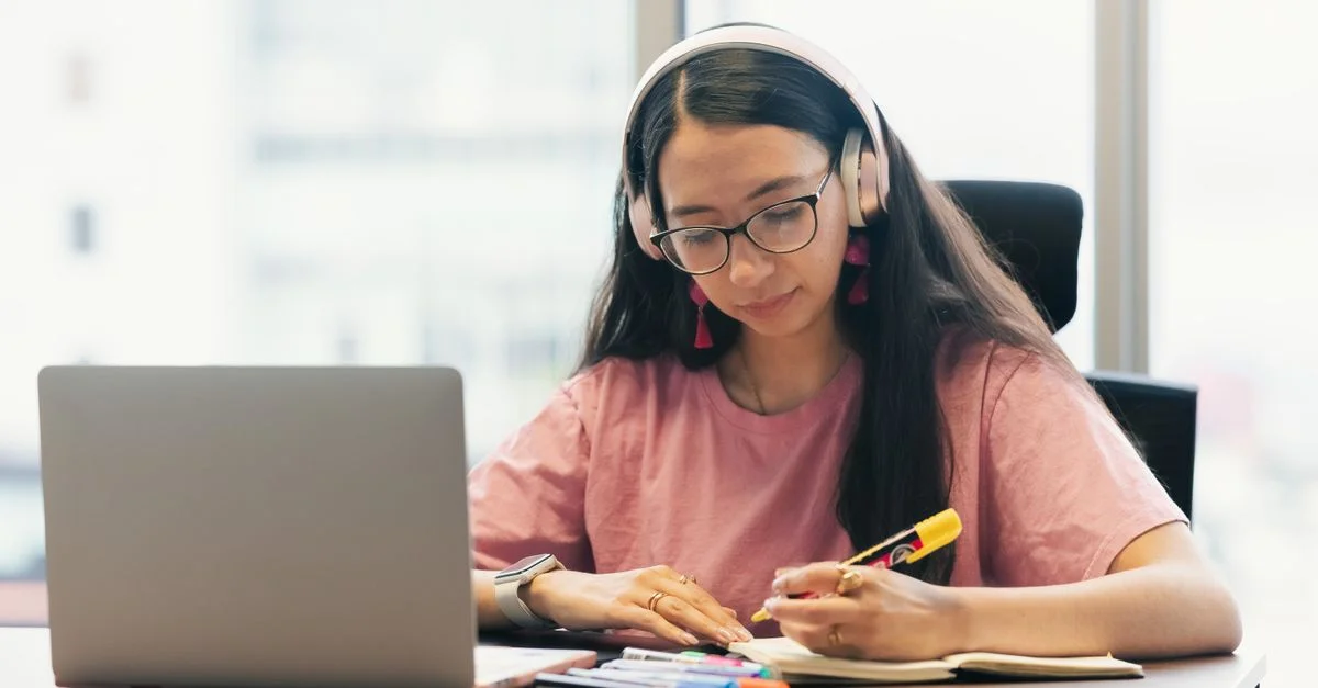 Femme jeune portant des écouteurs étudiant à un bureau avec un ordinateur portable et un cahier.