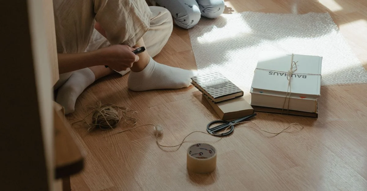Une scène cosy d'emballage de livres dans du film à bulles sur un sol en bois éclairé par le soleil, avec des outils et des matériaux autour.