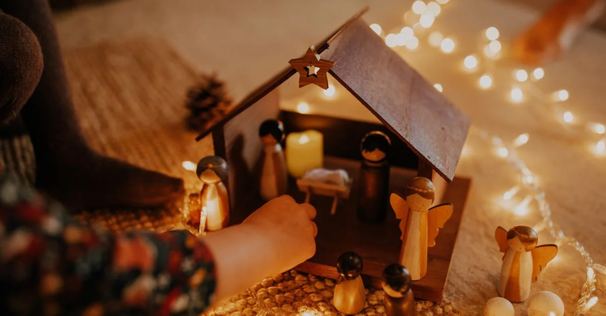 Un enfant arrange une crèche en bois entourée de lumières de Noël festives à l'intérieur.