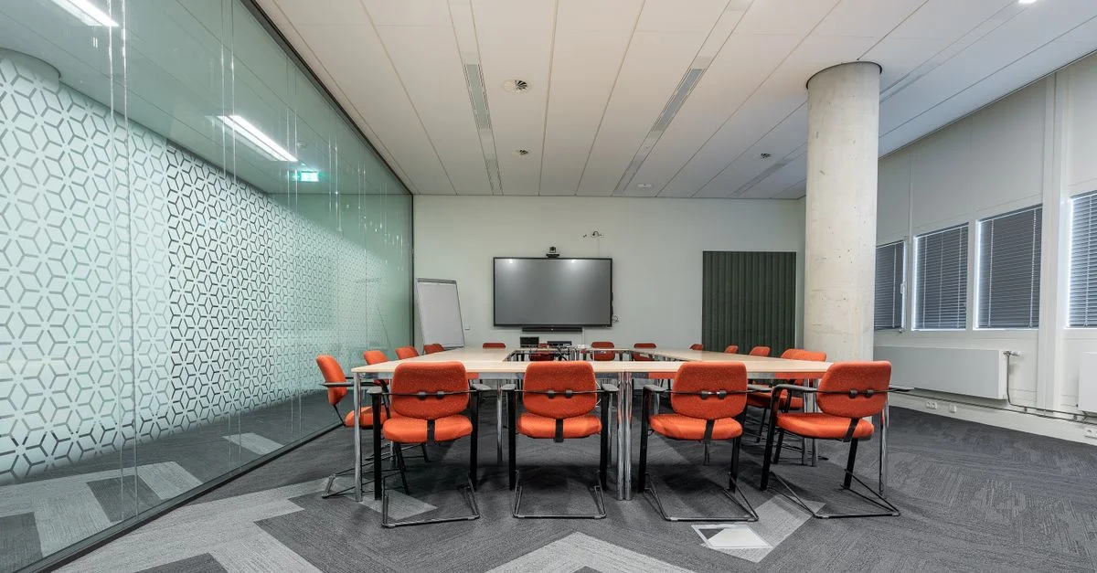 Salle de conférence spacieuse avec une grande table de réunion, des chaises rouges et des murs en verre pour un éclairage naturel.