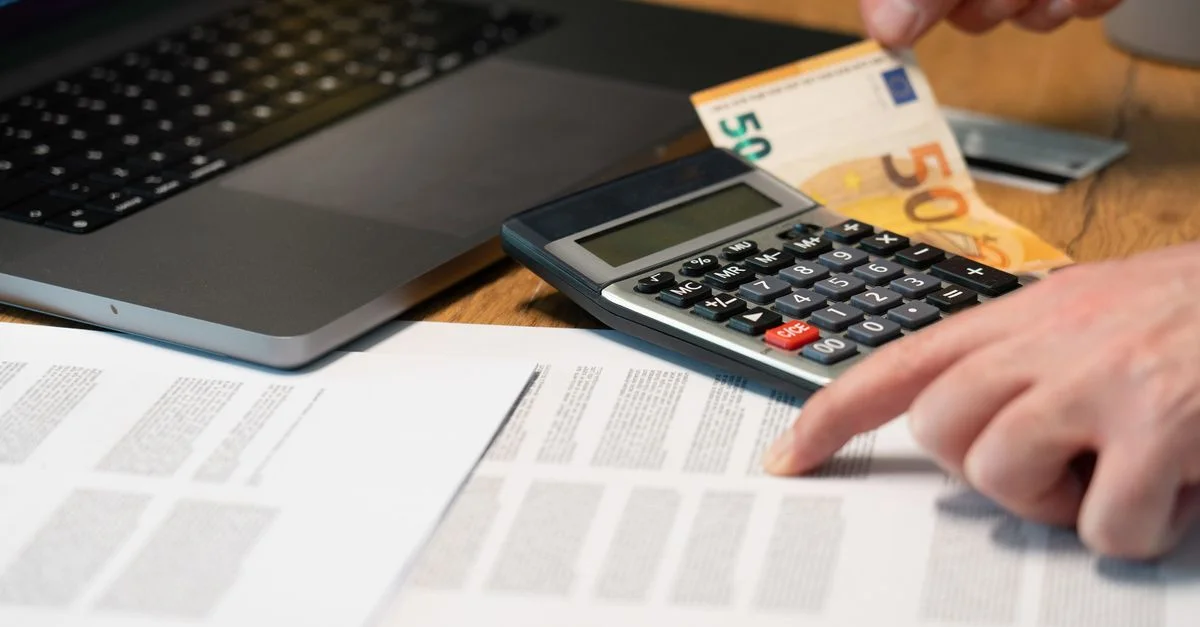 Gros plan des mains utilisant une calculatrice avec des billets en euros et un ordinateur portable sur un bureau.