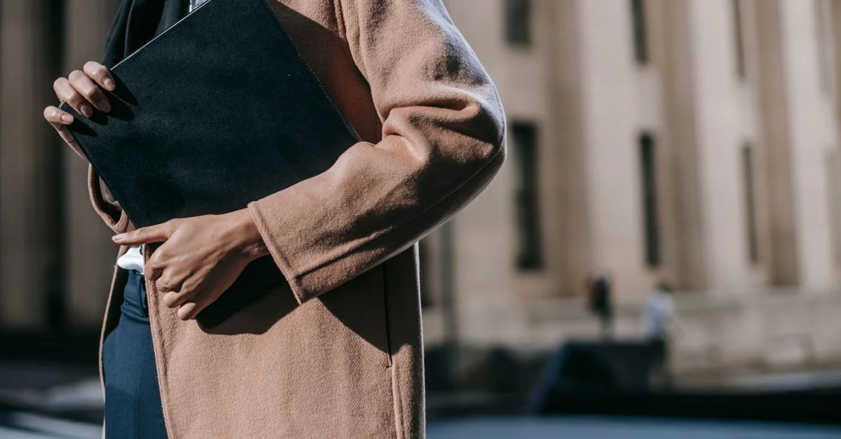 Une femme élégante en manteau tenant un dossier avec un bâtiment flou en arrière-plan, incarnant l'élégance professionnelle.