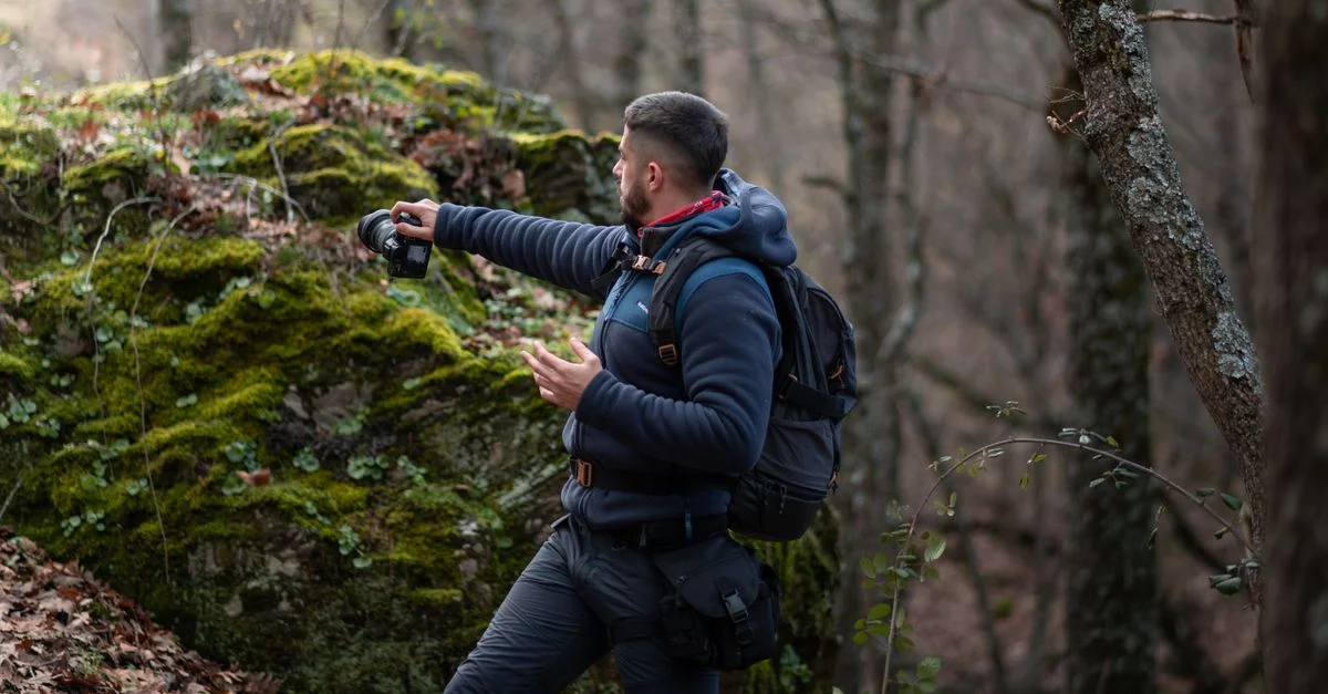 Un homme se tient dans une forêt couverte de mousse, capturant la nature avec son appareil photo. Scène de photographie en plein air aventureuse.