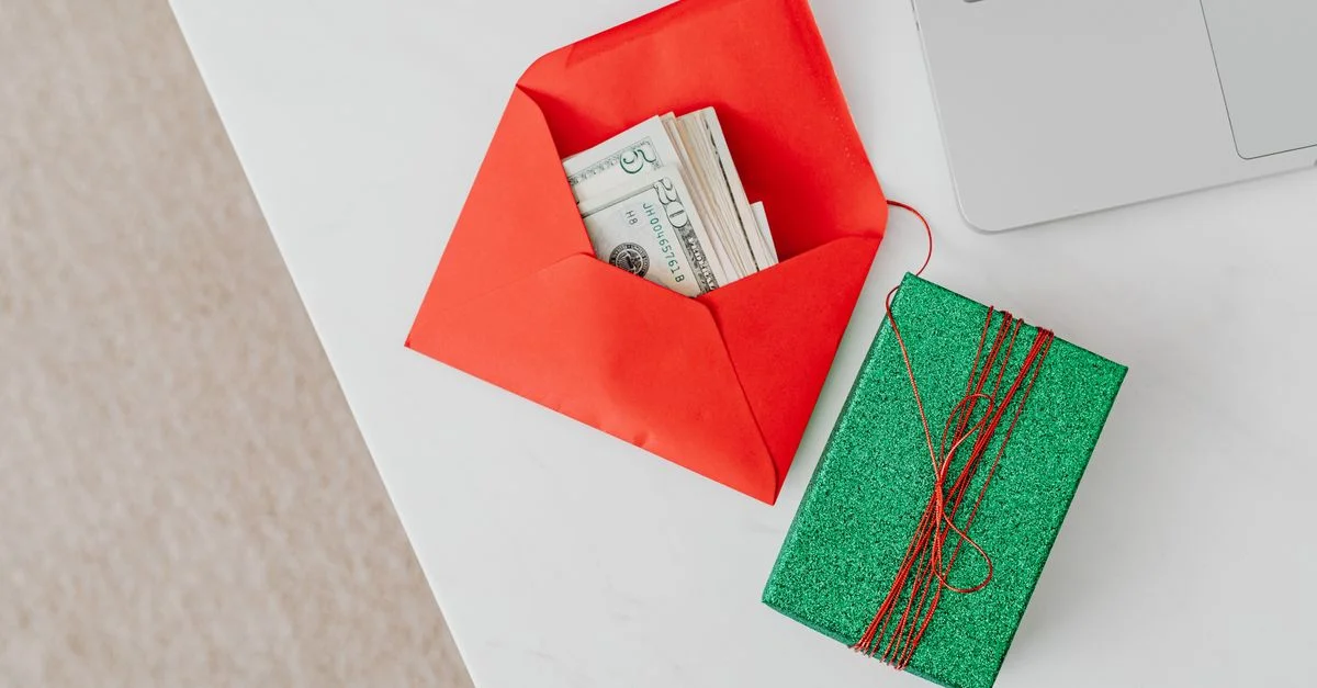 Une boîte-cadeau verte et une enveloppe rouge avec de l'argent sur un bureau à côté d'un ordinateur portable, soulignant des thèmes de vacances ou financiers.
