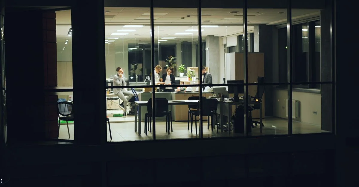 Des collègues collaborent dans un bureau vitré la nuit, concentrés sur leurs tâches.