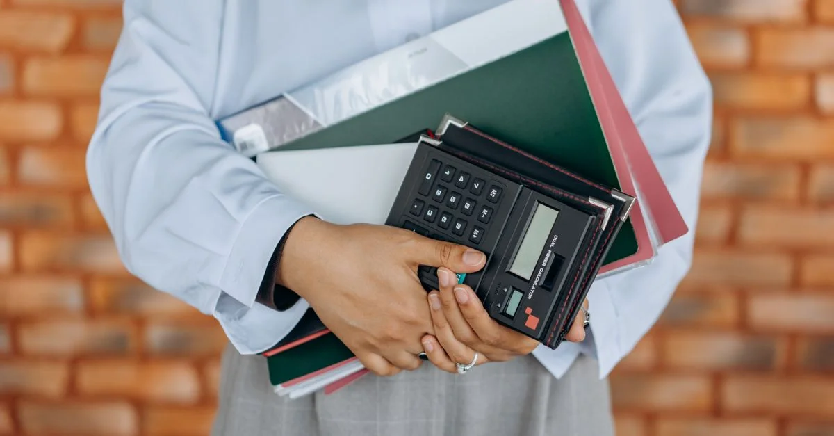 Gros plan sur une personne tenant une calculatrice et des dossiers sur fond de mur en briques.