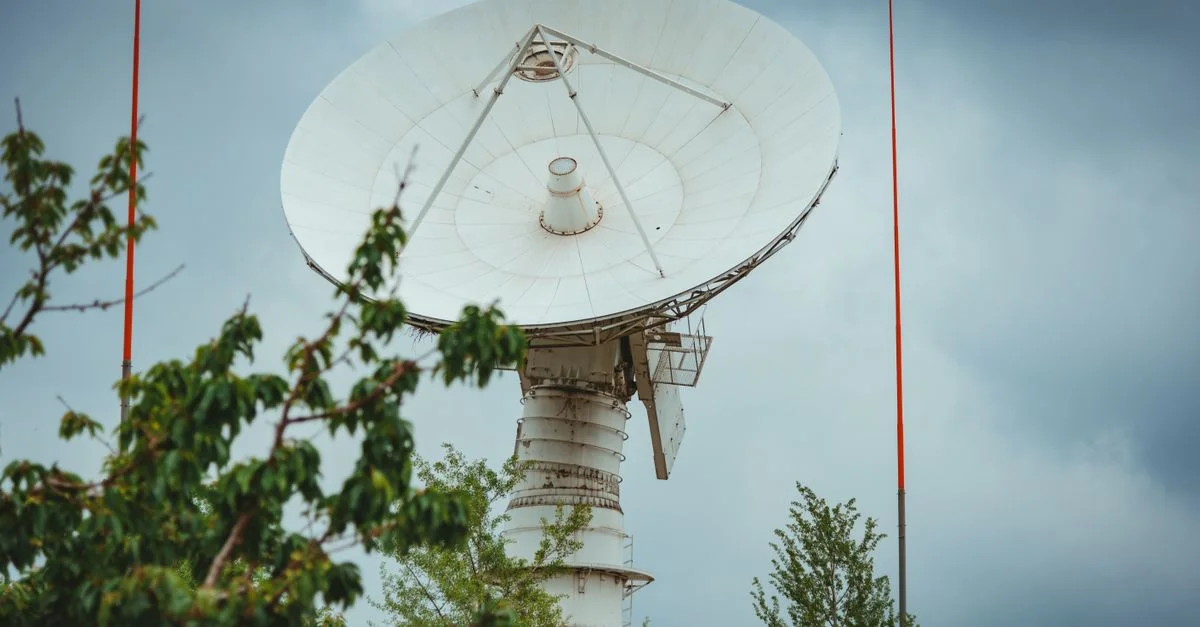 Une grande antenne satellite parabolique avec un ciel nuageux en arrière-plan, entourée de verdure.
