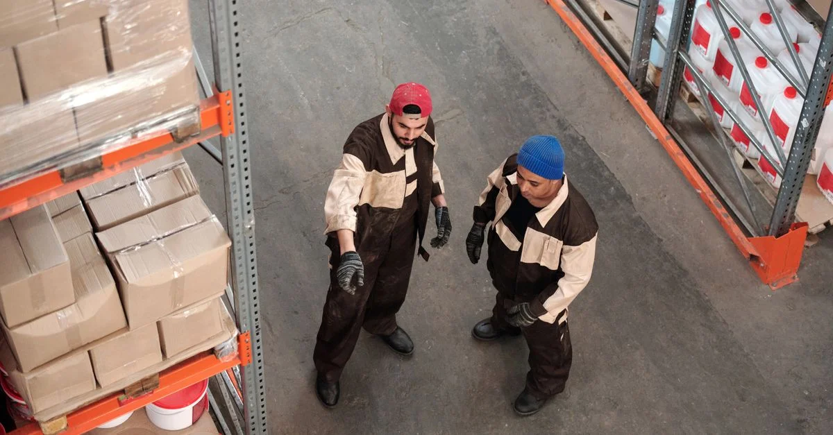 Deux hommes adultes dans un entrepôt discutant de la logistique de stockage, entourés d'étagères de colis.