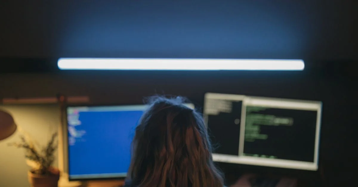 Un développeur logiciel travaillant dans une pièce faiblement éclairée avec deux écrans affichant du code, illuminée par une lumière blanche.