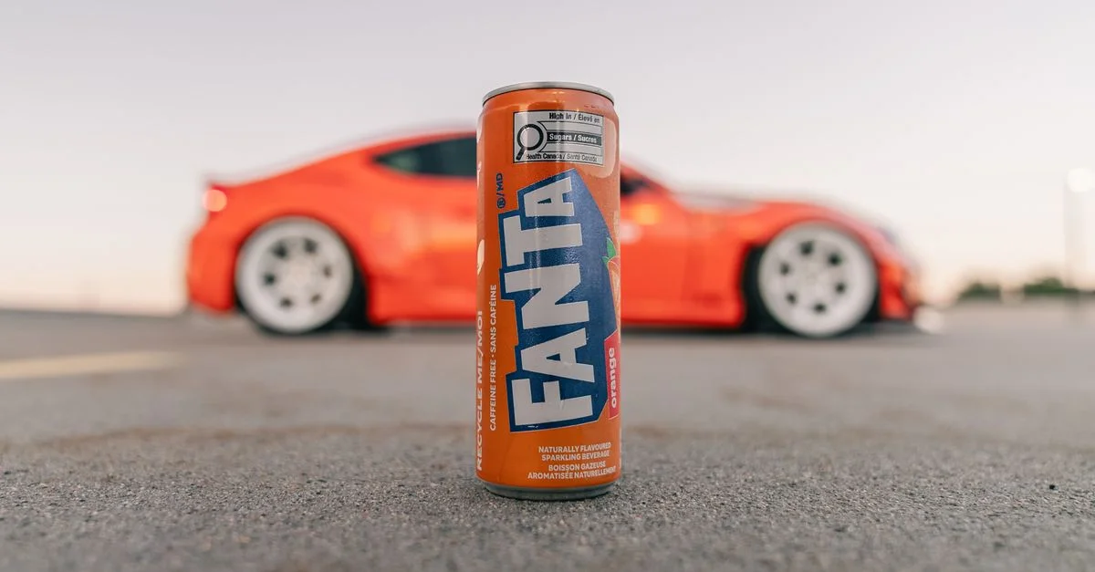 Une canette de Fanta aux couleurs vives se détache sur un fond de voiture de sport rouge, dans un parking désert au crépuscule.