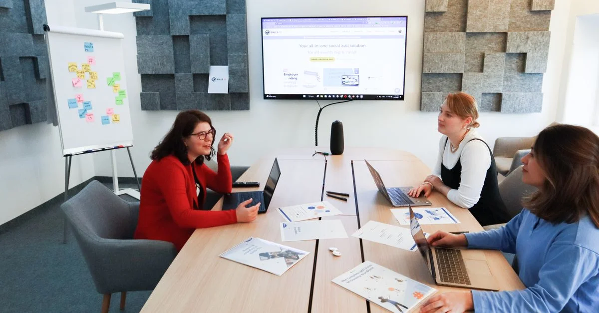 Trois femmes dans un bureau moderne engagées dans une discussion de stratégie commerciale avec des ordinateurs portables et un écran de présentation.