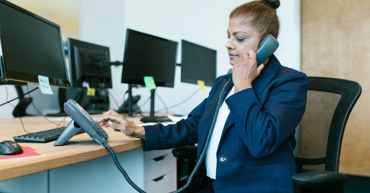 Femme d'affaires multitâche au bureau, utilisant un téléphone et un ordinateur, vêtu d'une tenue formelle.