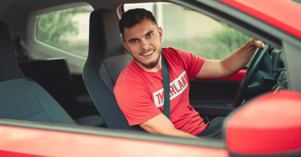Homme souriant conduisant une voiture rouge tout en portant une ceinture de sécurité, mettant en avant la sécurité et la joie.