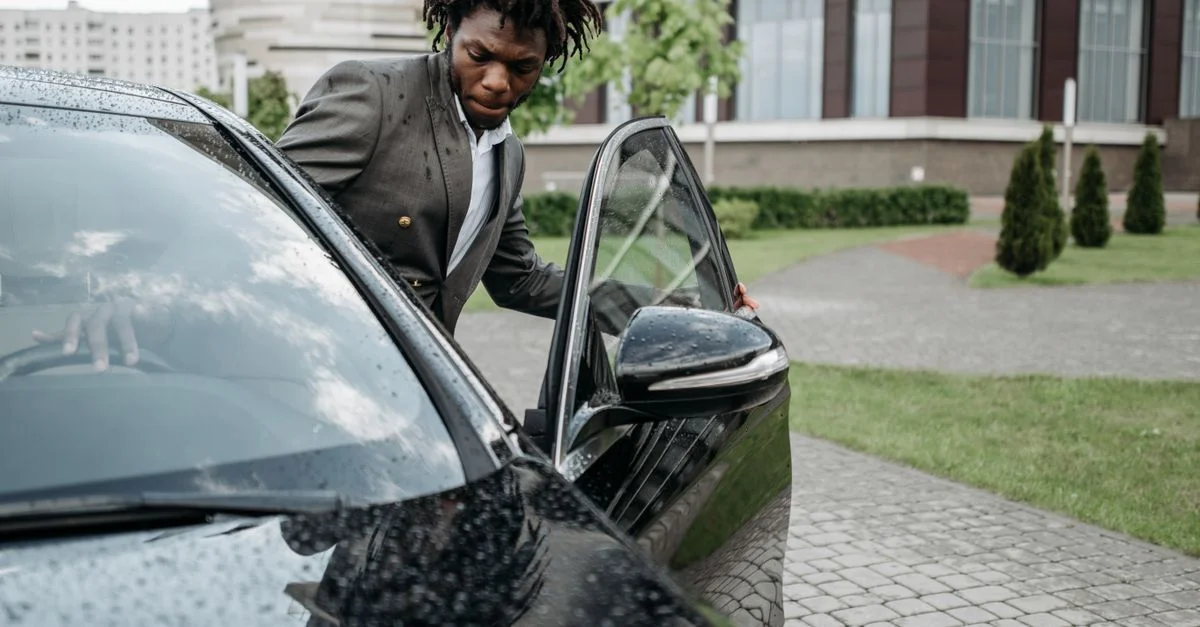 Un homme afro-américain en costume gris sort d'une voiture noire devant un immeuble de bureaux moderne par une journée pluvieuse.