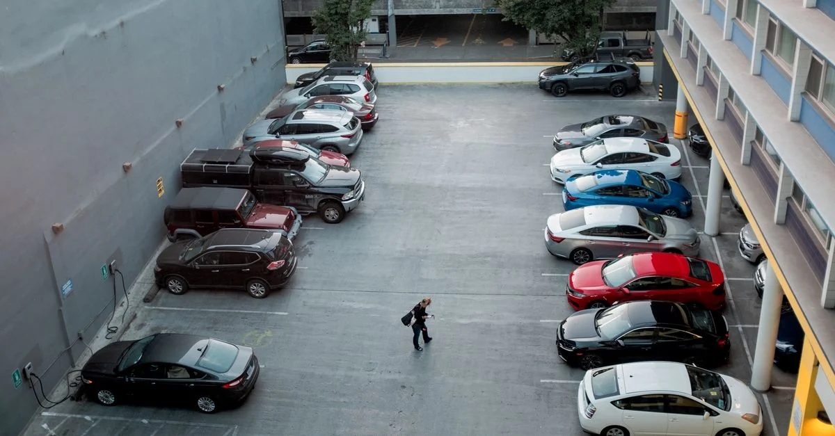 Un homme marche dans un parking de ville rempli de diverses voitures, offrant une scène urbaine animée.