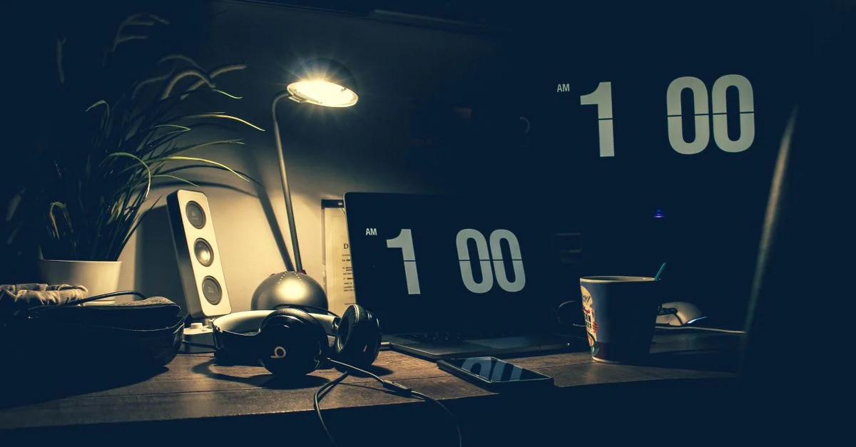 Un bureau moderne faiblement éclairé avec un ordinateur portable, des écouteurs et des enceintes à 1h00 du matin.