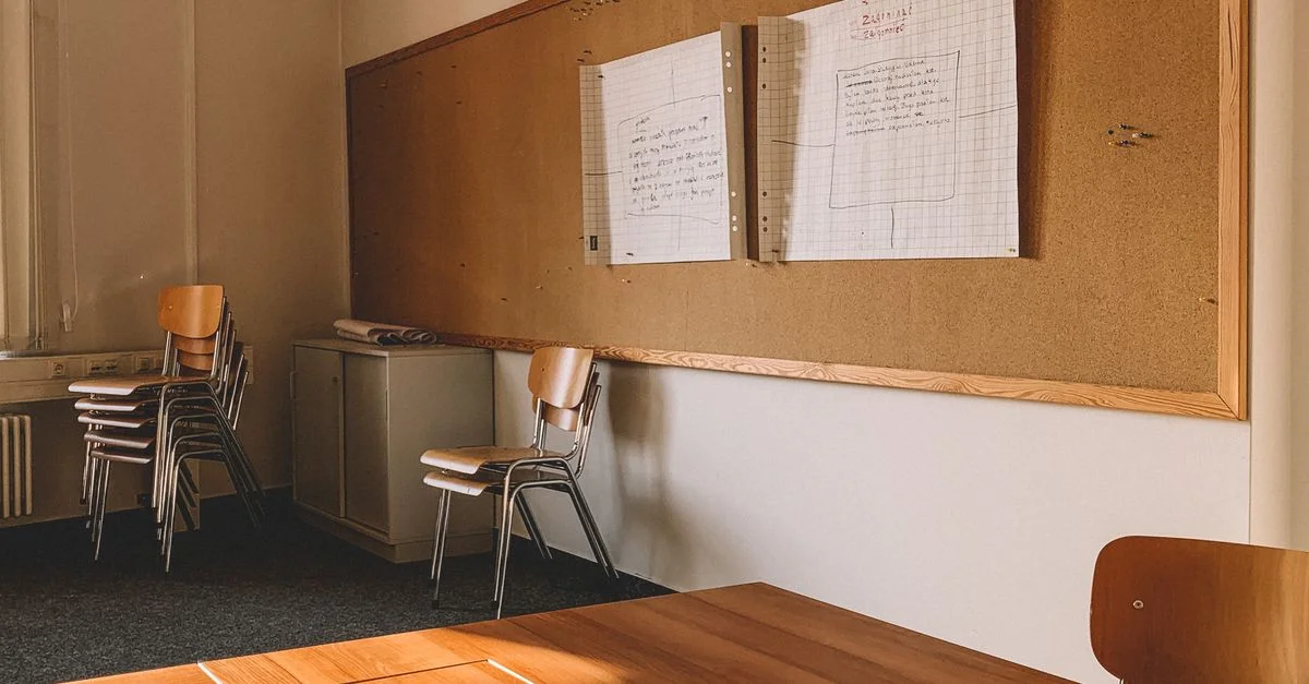 Une salle de classe vide avec des chaises empilées, des tables et un tableau en liège sur le mur.