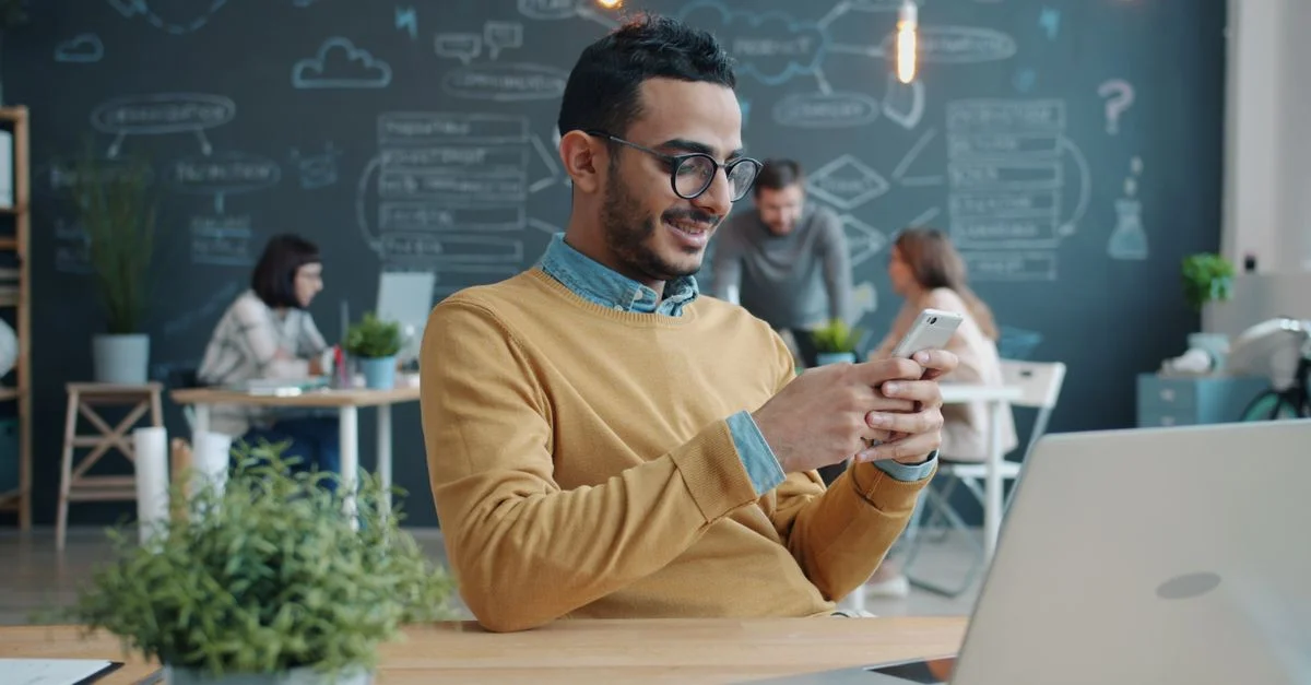 Un homme souriant utilise son smartphone dans un bureau moderne et décontracté, ses collègues travaillant en arrière-plan.