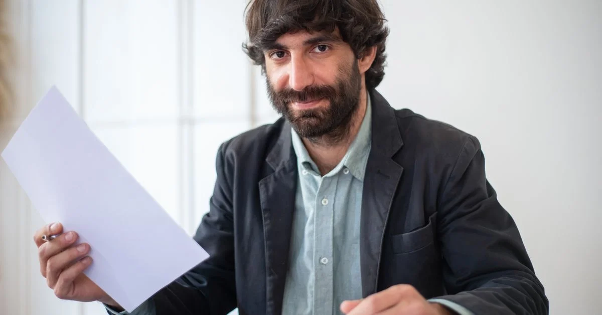 Un homme barbu en veste noire tenant des documents, symbolisant un environnement de bureau professionnel.
