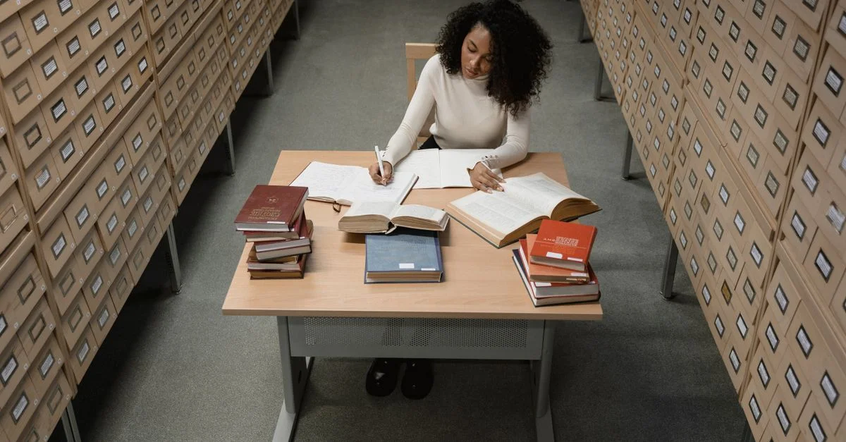 Une femme afro-américaine étudiant à un bureau en bois entouré de tiroirs dans une salle d'archives.