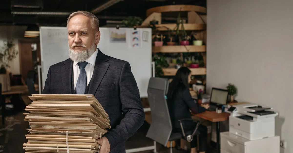 Homme âgé en costume tenant une grande pile de documents dans un bureau moderne.